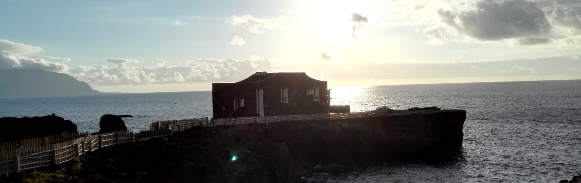 Reddish-brown building perched on dark volcanic rocks jutting into the Atlantic Ocean, with wooden walkway and distant cliffs under cloudy sky.
