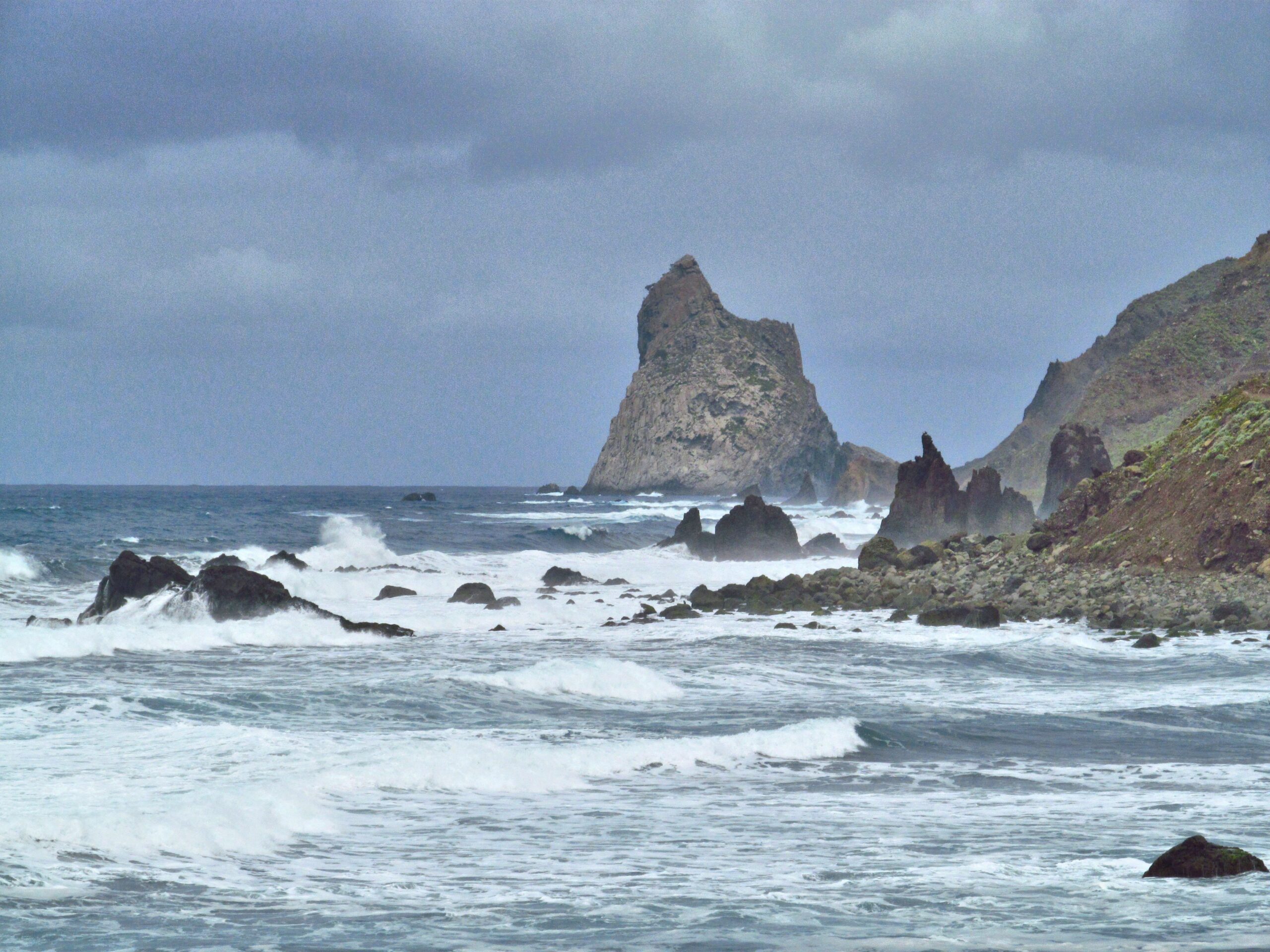 Tall volcanic rock formation rising from the sea with waves crashing against dark rocks and cliffs in the background under cloudy sky
