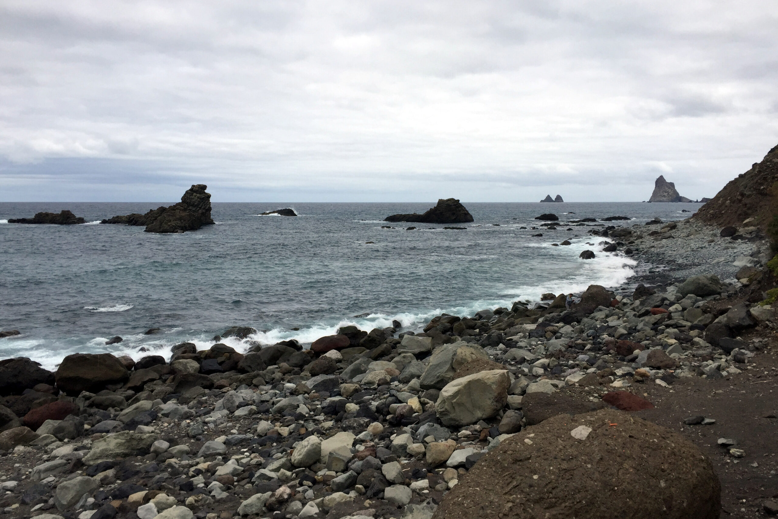 Rocky coastline with dark volcanic boulders and pebbles along the shore, white-capped waves, and dark volcanic rock formations rising from the sea under overcast sky