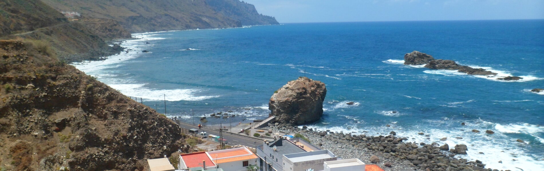 Colorful houses with red and green roofs on a rocky coastal slope overlooking blue Atlantic waters, jagged sea stacks, and dramatic green mountains.