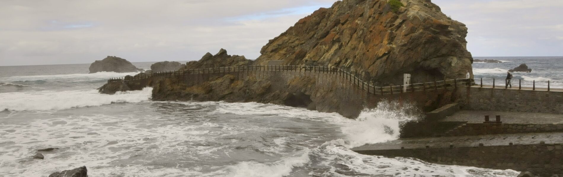 Rocky coastal outcrop with a white cross at its peak, wooden railings along a sandy beach, white-capped waves, and overcast sky
