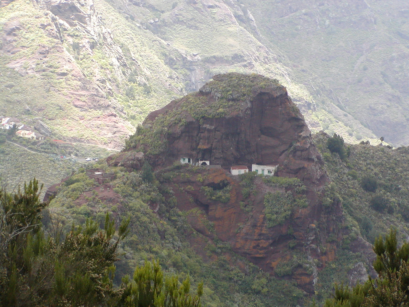 Isolated white buildings perched on a steep rocky outcrop covered in green vegetation, surrounded by deep ravines and distant mountain slopes.