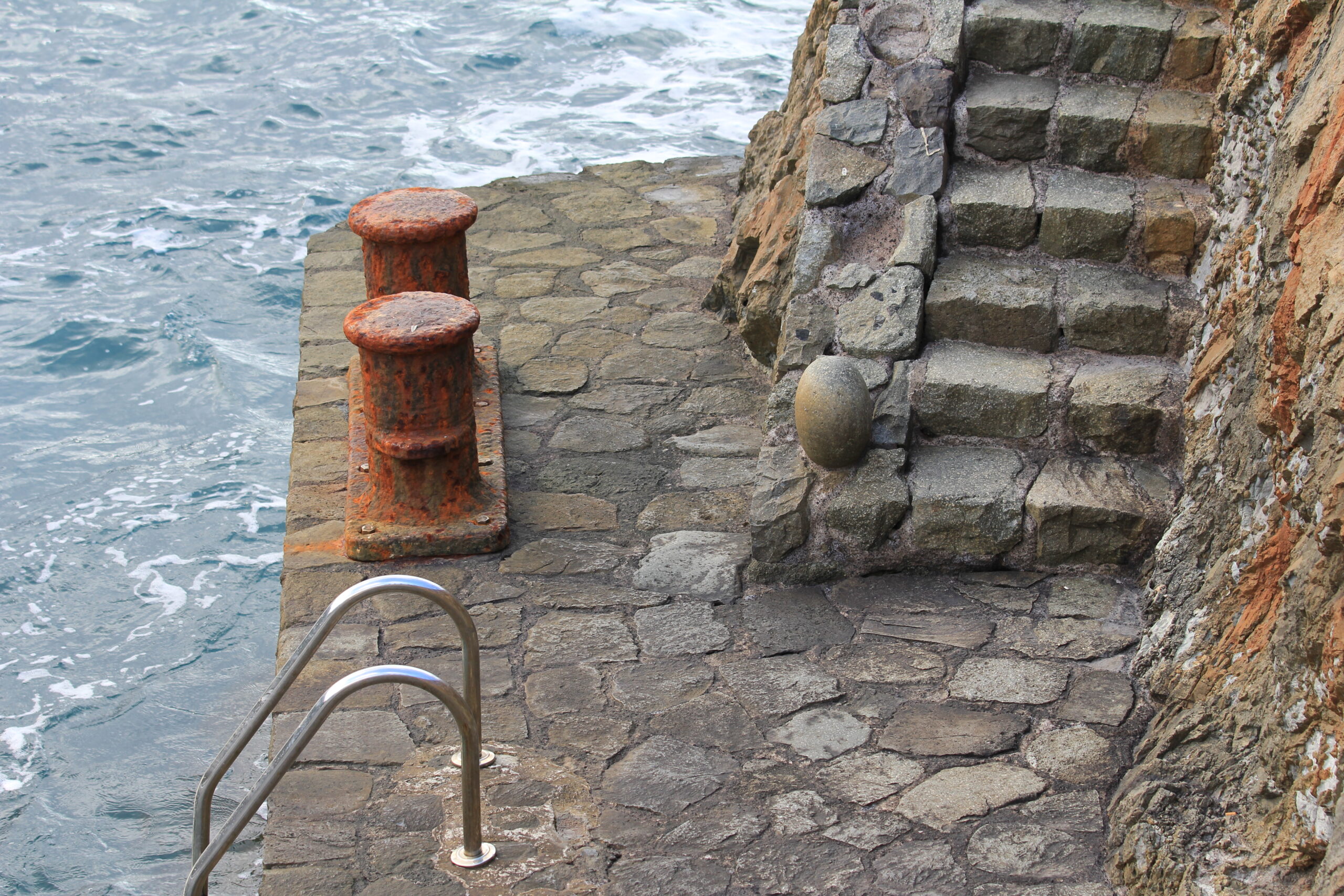 Weathered stone steps descend to a rocky coastal platform with rusty mooring bollard and metal handrail, Atlantic waves visible beyond.