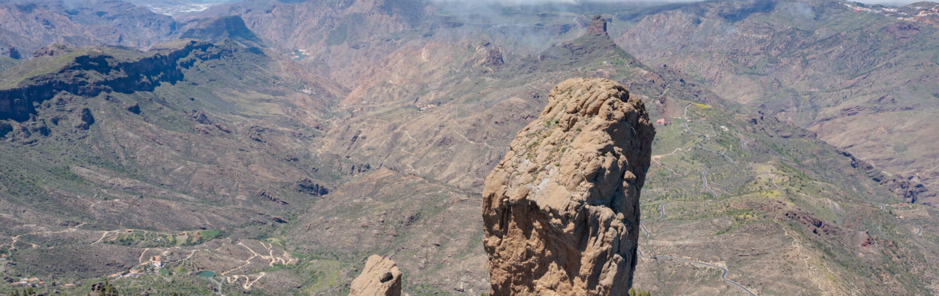 Distinctive tall rock pinnacle rising from a forested mountainside, with deep valleys and distant volcanic peaks visible beyond a cloud layer.