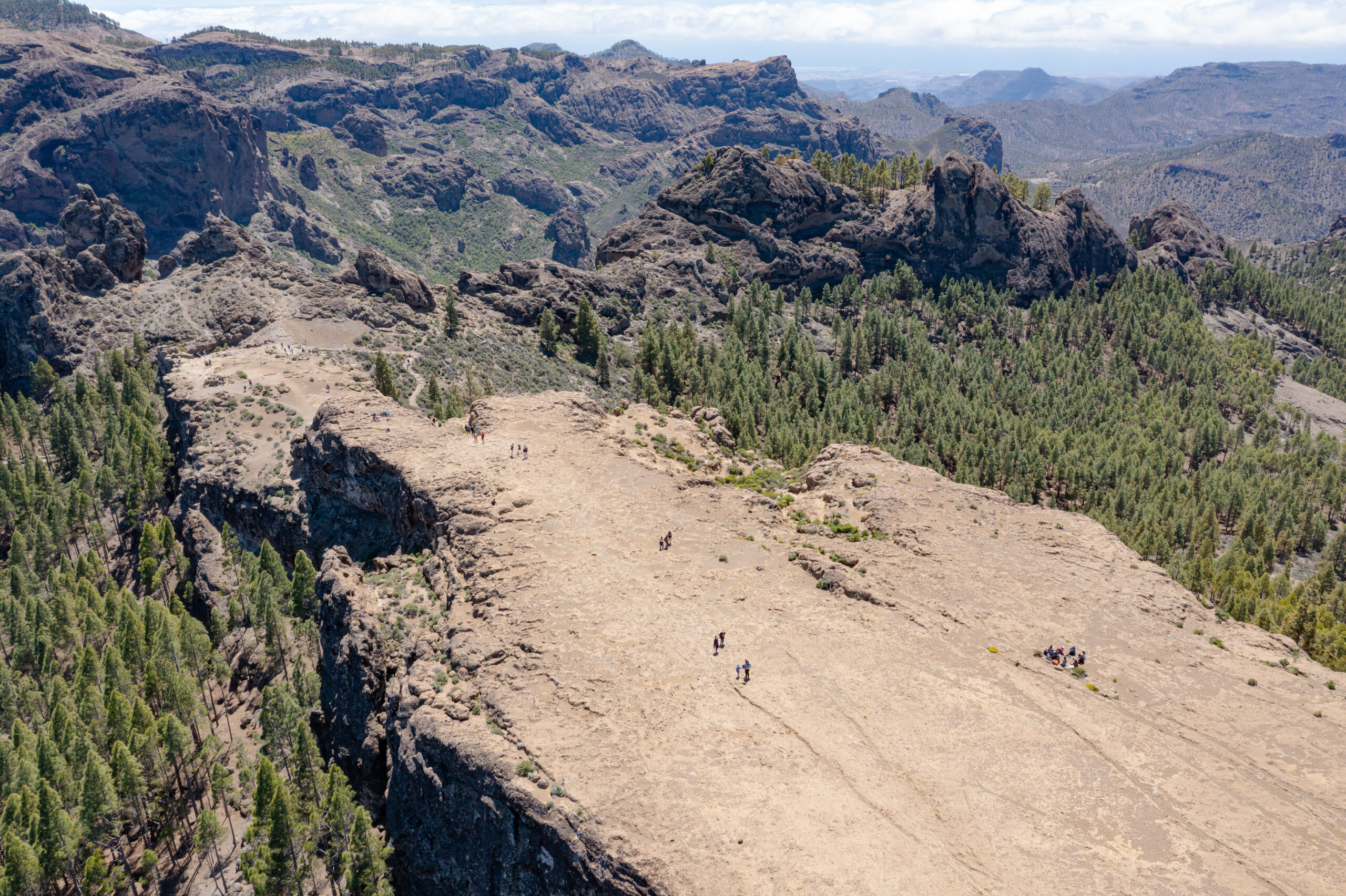 Roque Nublo's high-altitude plateau offers sweeping views of volcanic rock formations and pine forest, with hikers scattered across the sandy summit.