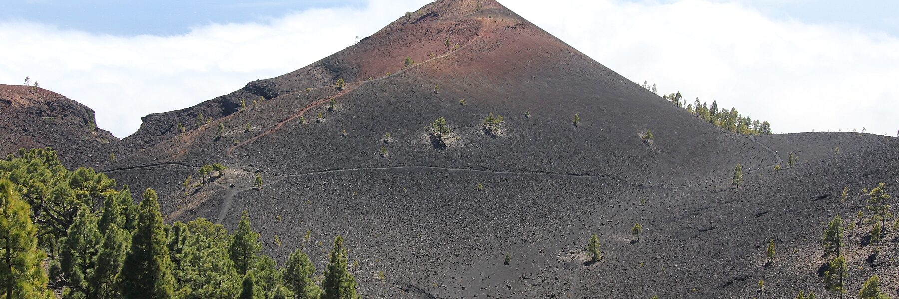 Dark volcanic cone with reddish-brown summit rising above sparse pine forest on black lava fields under partly cloudy sky