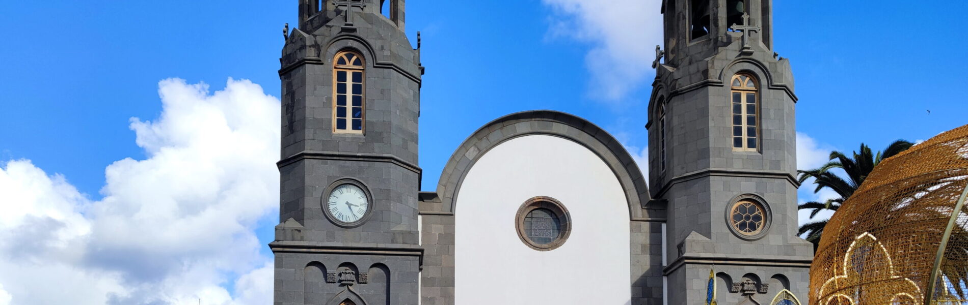 Stone church with white facade and two tall spires topped with crosses, set against blue sky with clouds