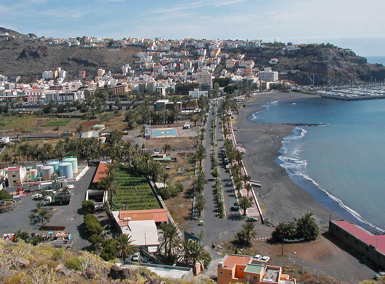 San Sebastián's beachfront curves along dark volcanic sand, lined with palms and backed by colorful buildings cascading up the steep hillside.