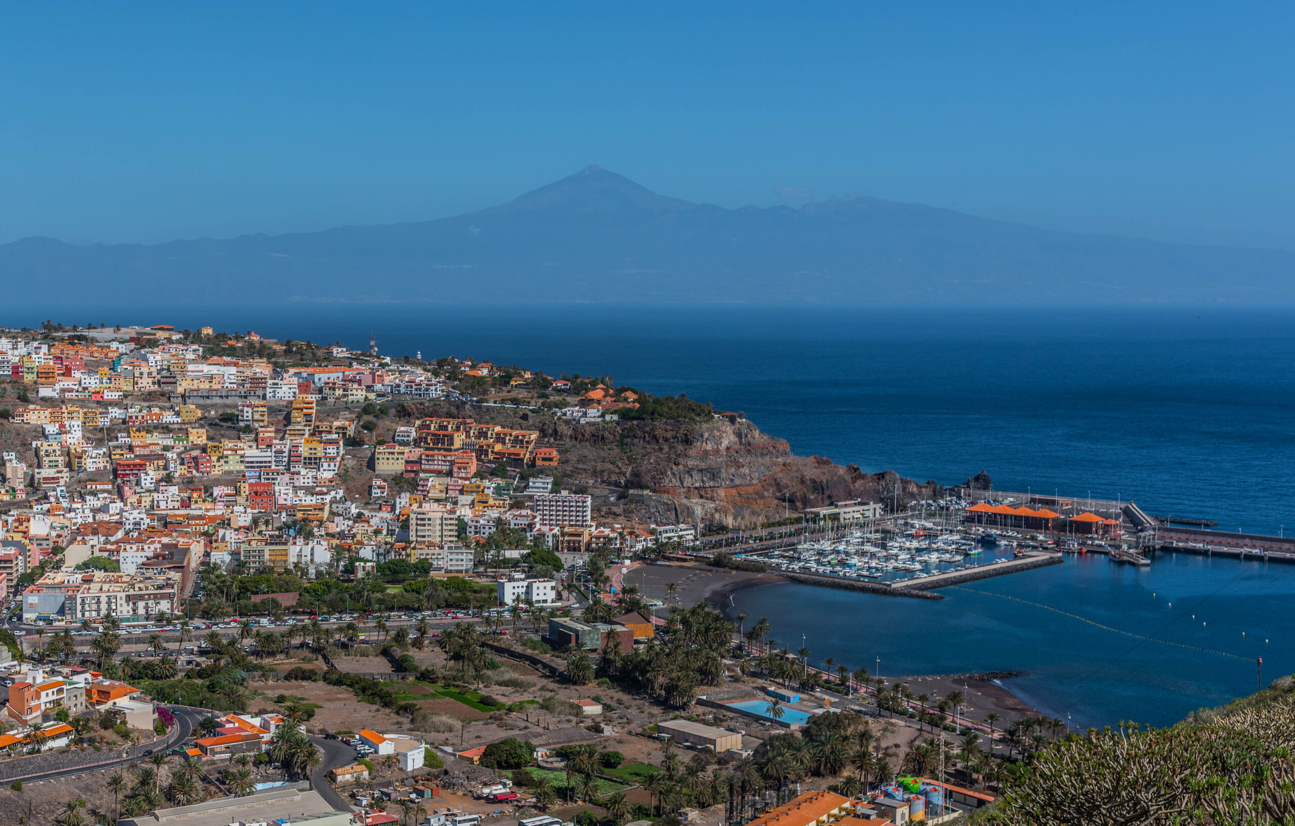 San Sebastián's waterfront settlement spreads across volcanic slopes overlooking a sheltered marina, with Tenerife's Mount Teide visible on the distant horizon.