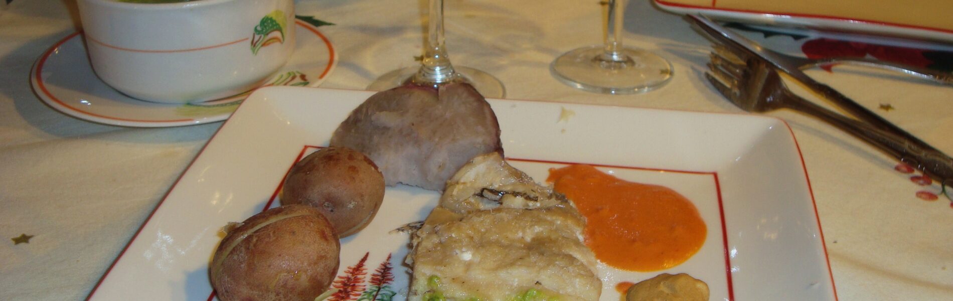 Plated traditional Canarian dish with fish, potatoes, vegetables, and sauces on white plate with red border, accompanied by wine glasses and soup bowl