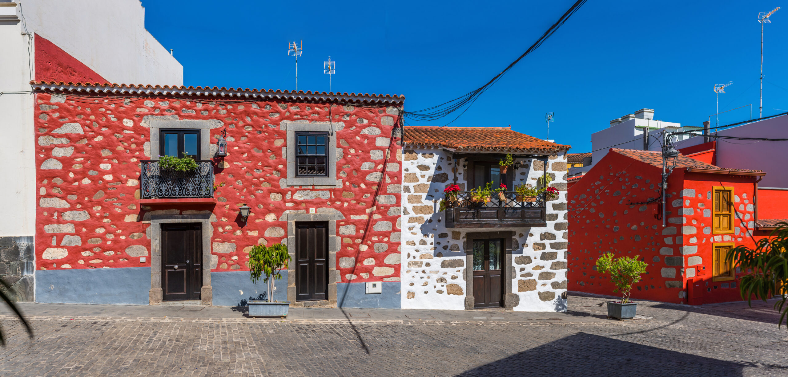 Vibrant traditional architecture in Santa Brígida, featuring red-painted stone buildings with wooden balconies and doors arranged around a quiet village square.