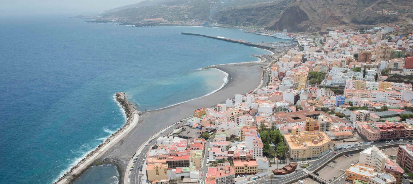Aerial view of Santa Cruz de La Palma with curved black-sand beach, harbor breakwater, and volcanic hillside under blue sky