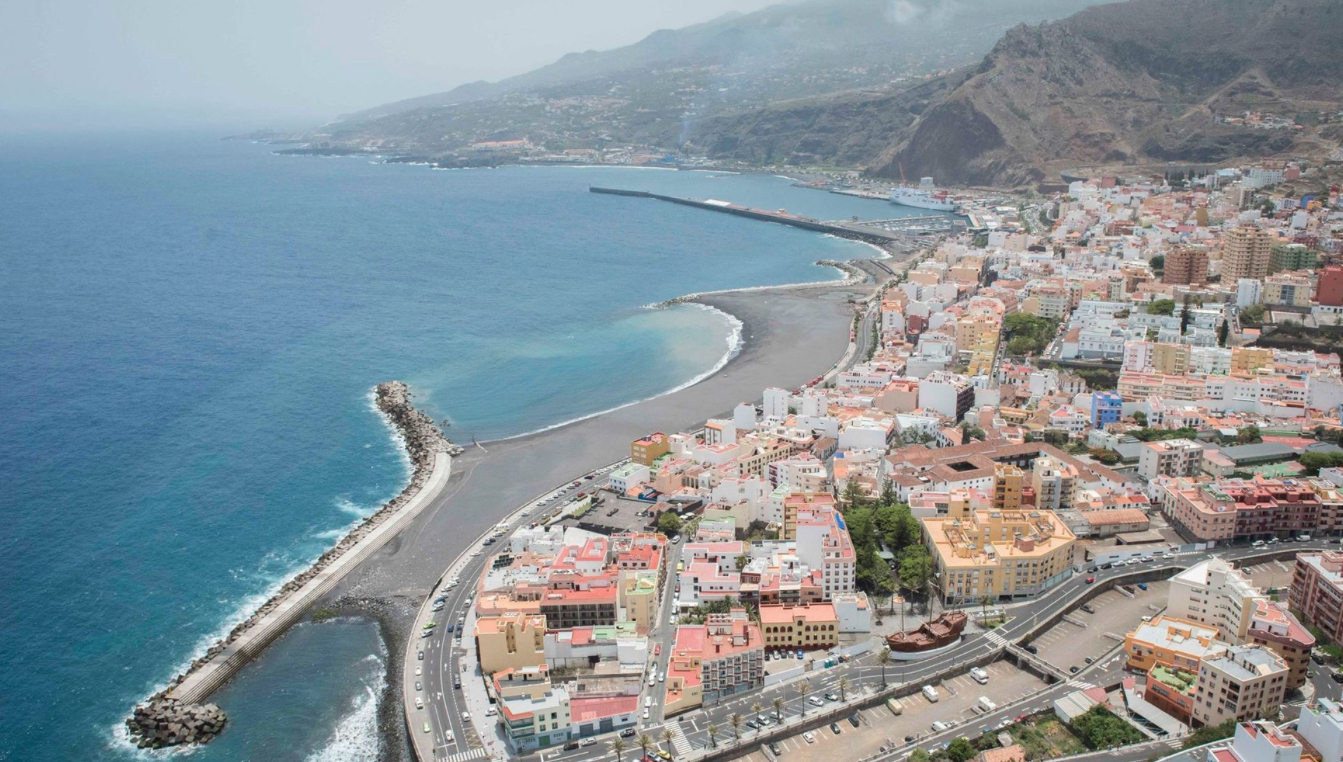 Santa Cruz de La Palma's distinctive crescent beach curves along the coast, sheltered by a long breakwater, with the town's colorful buildings rising up the volcanic slopes behind.
