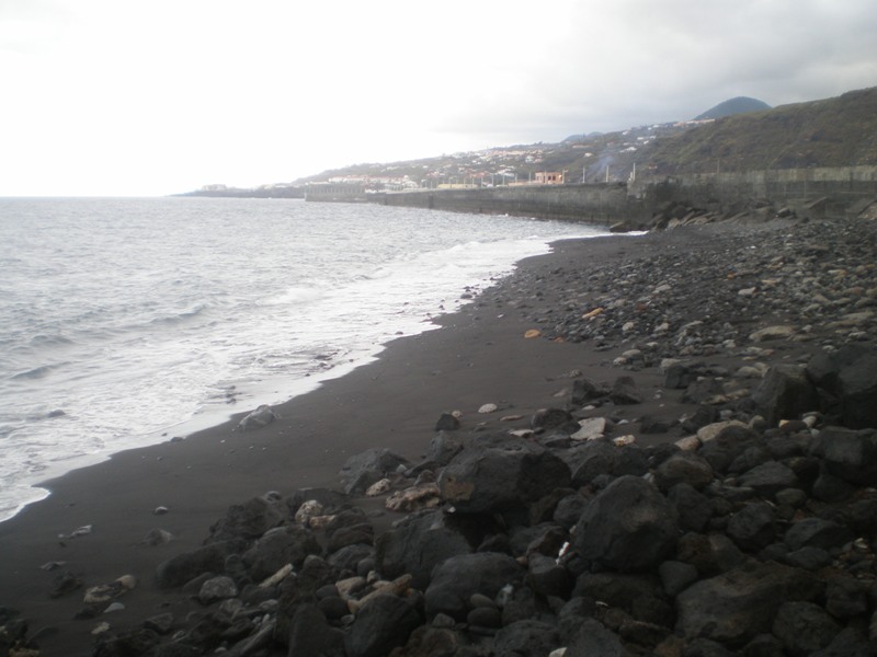 The dark volcanic sand and rocky shoreline of Santa Cruz de La Palma's coast, with the town cascading up the hillside behind.