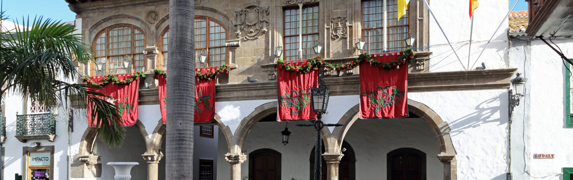 Colonial-era stone building with arched portico, decorative stonework, red flower garlands, and Spanish flags under clear blue sky with palm tree in foreground.