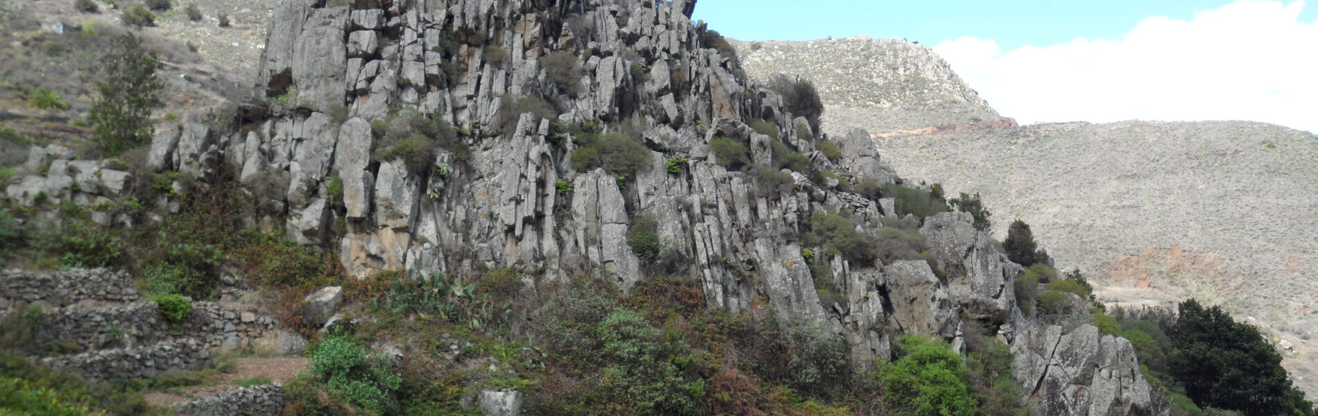 Dramatic columnar basalt rock formation rising above scrubland and vegetation on a hillside under clear blue sky
