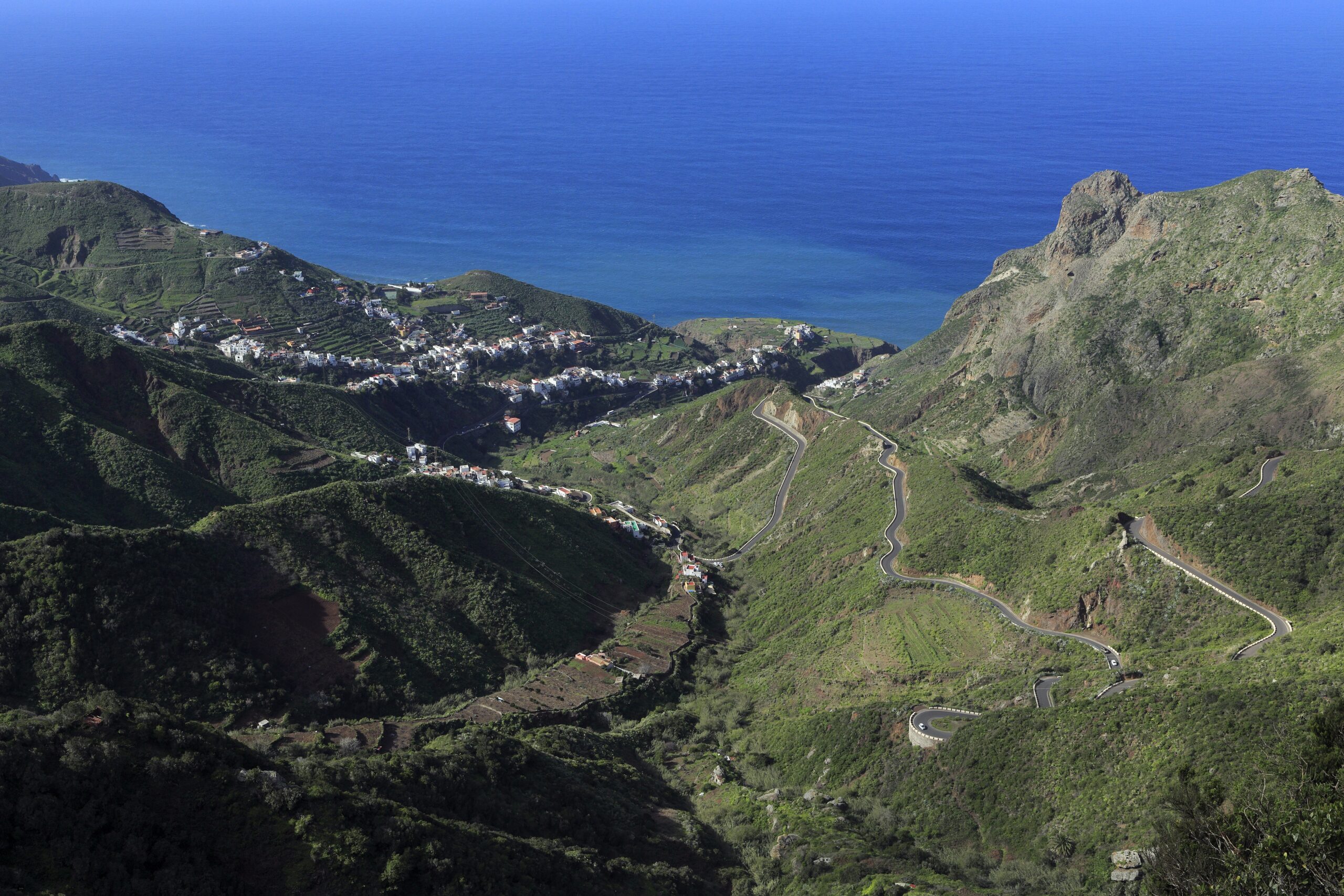 Aerial view of green mountainous terrain with a winding road, white buildings nestled in a valley, and deep blue ocean beyond