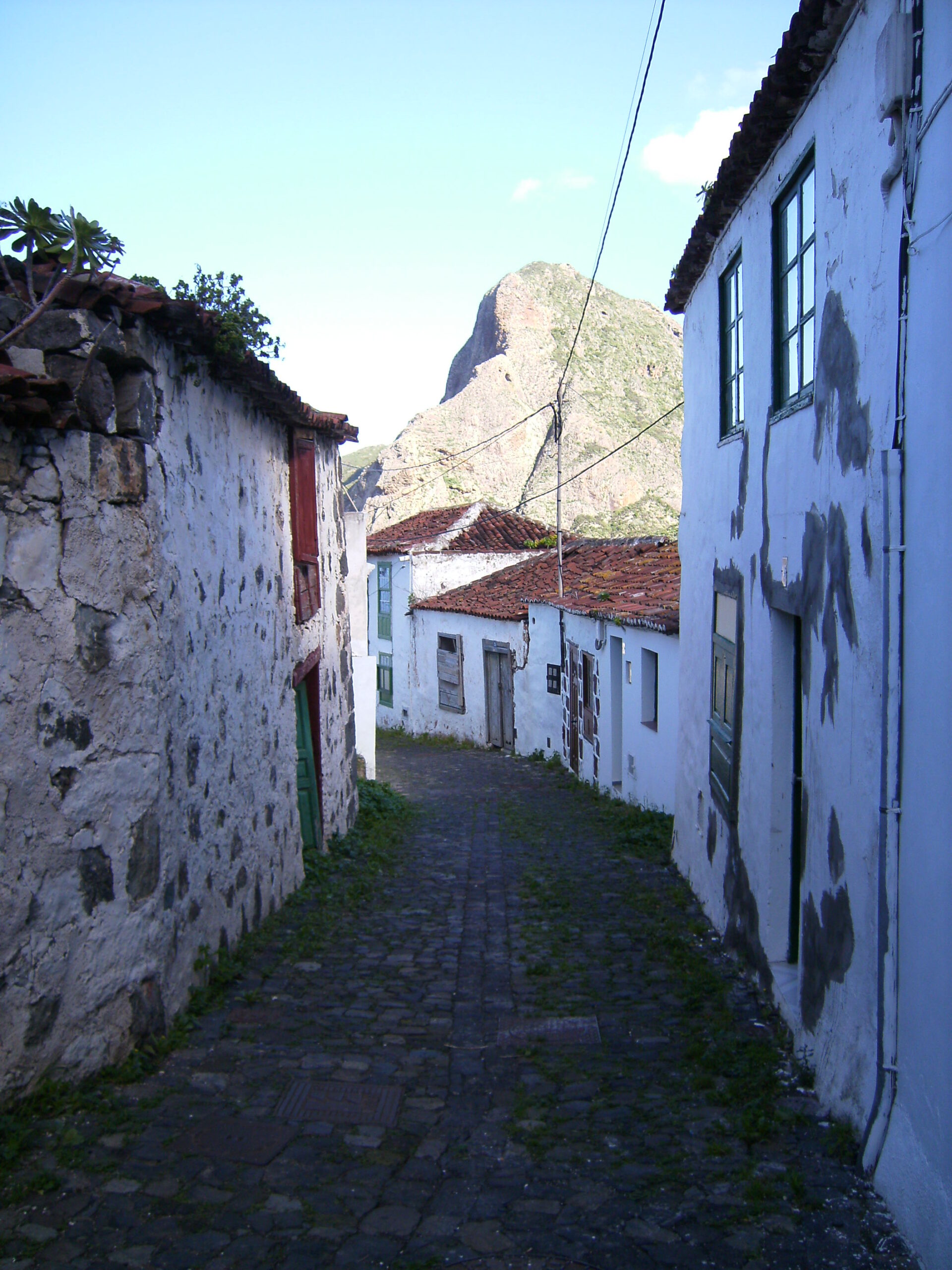 Narrow cobblestone alley between whitewashed houses with a dramatic rocky cliff rising in the background under clear sky