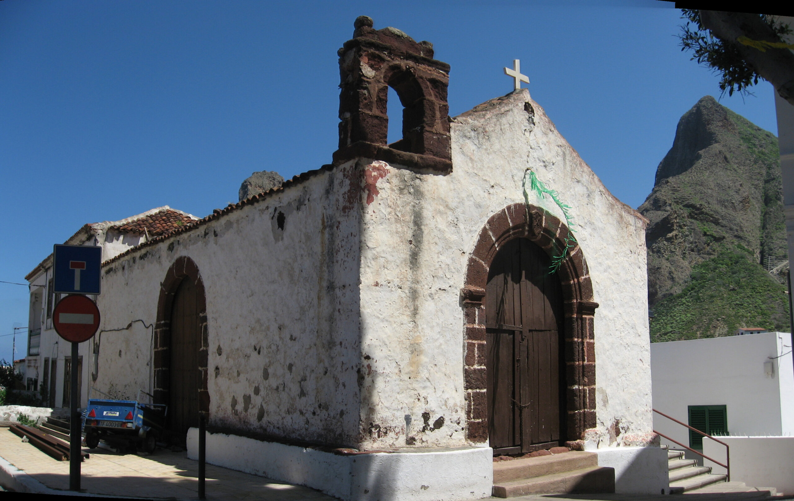 Whitewashed stone chapel with weathered brick bell tower, wooden arched door, white cross on roof, and steep volcanic rock formation behind it