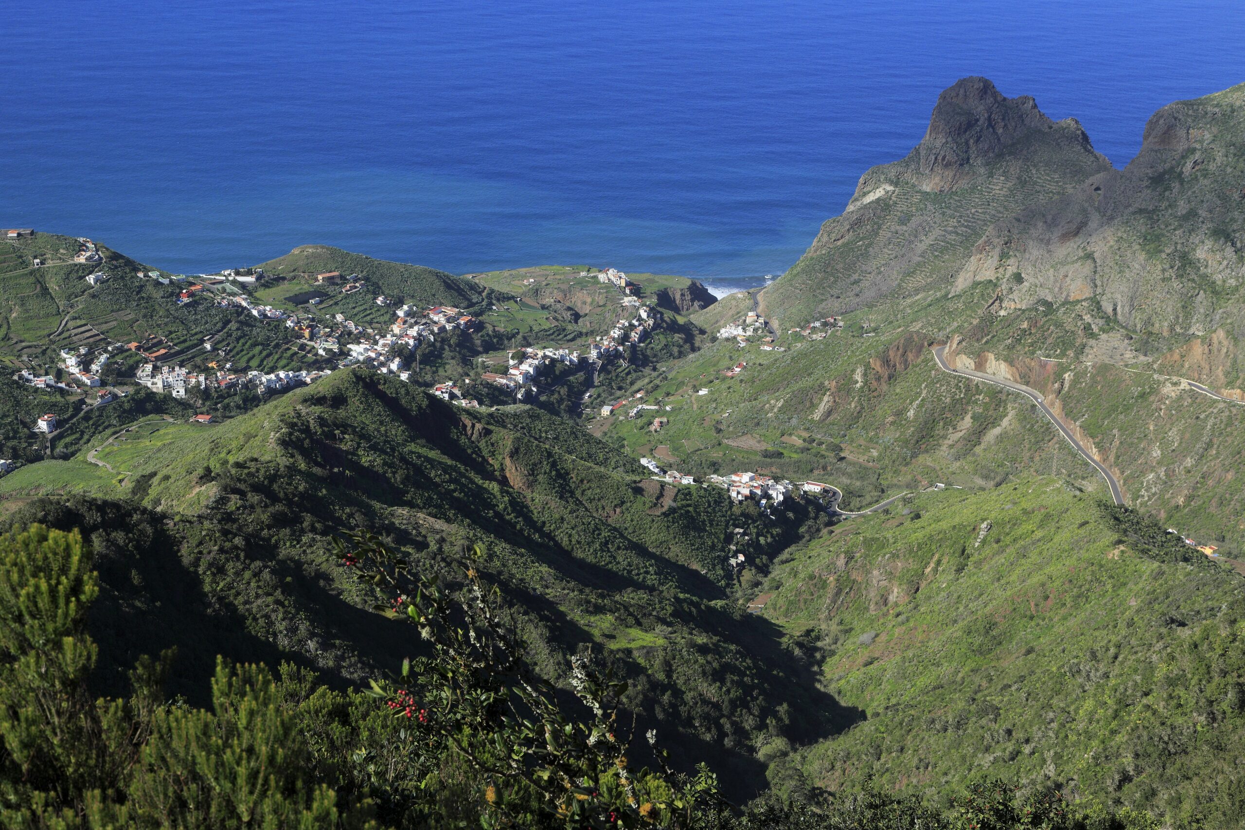 Aerial view of Taganana village nestled in green volcanic valleys with white buildings scattered across hillsides, a dramatic rocky peak, and the deep blue Atlantic Ocean beyond.