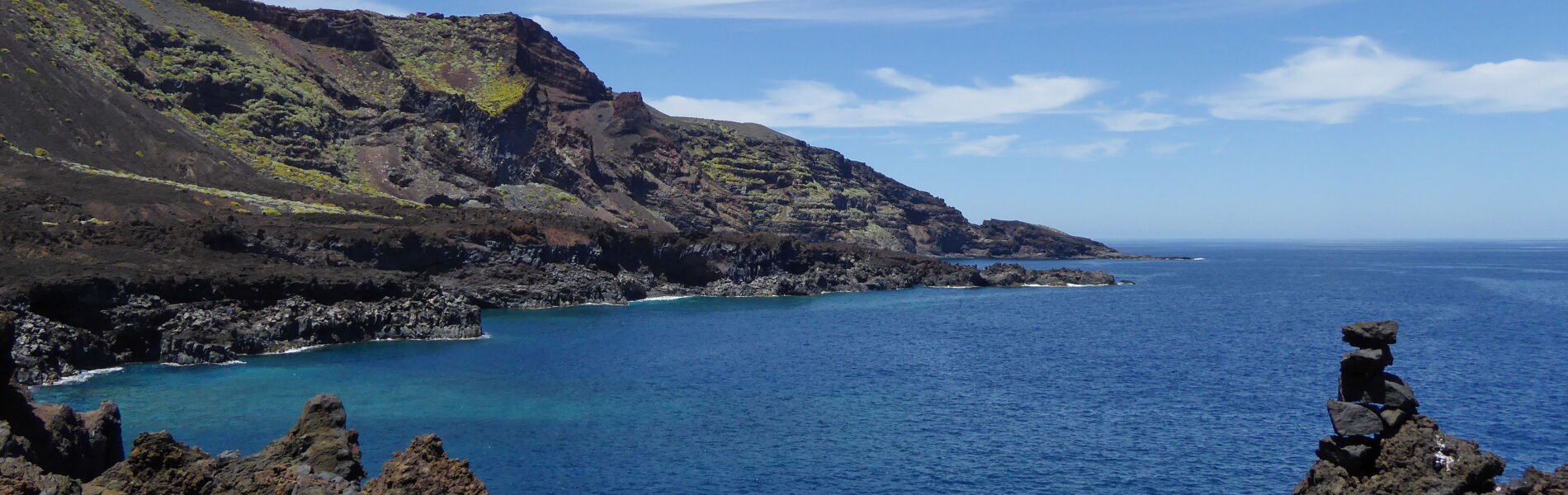 Turquoise cove surrounded by volcanic cliffs covered in moss-green vegetation, with dark rocky outcrops in the foreground and deep blue ocean beyond under clear sky.