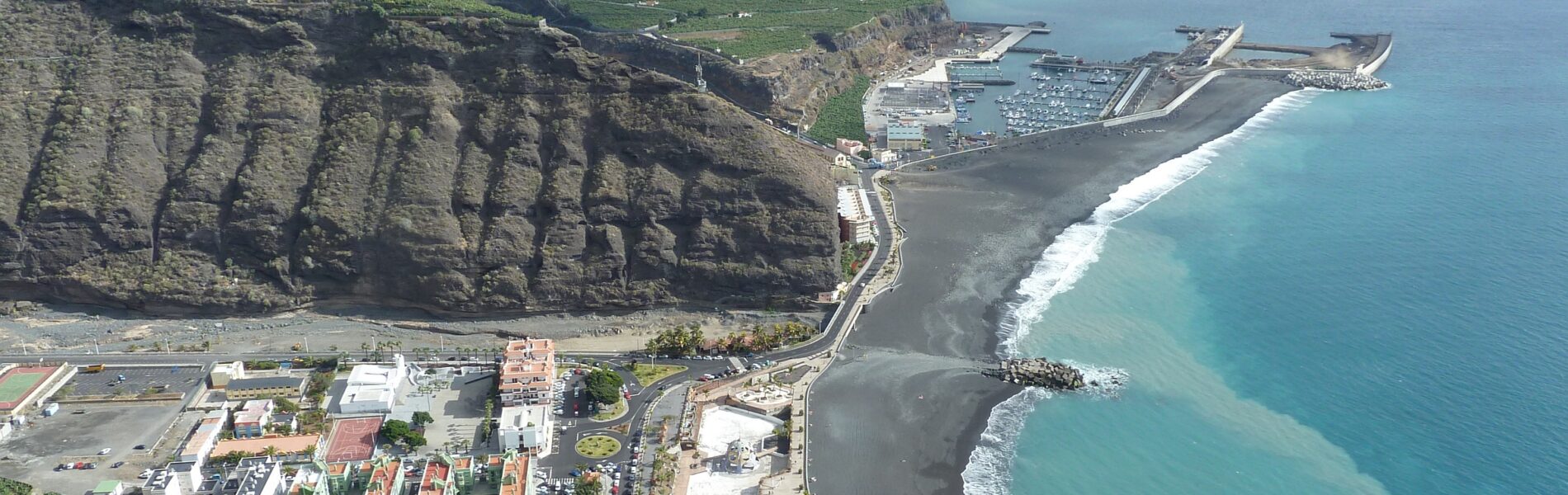 Aerial view of Tazacorte beach with dark volcanic sand, turquoise waters, harbor with ships, residential buildings on slopes, and steep basalt cliffs rising behind