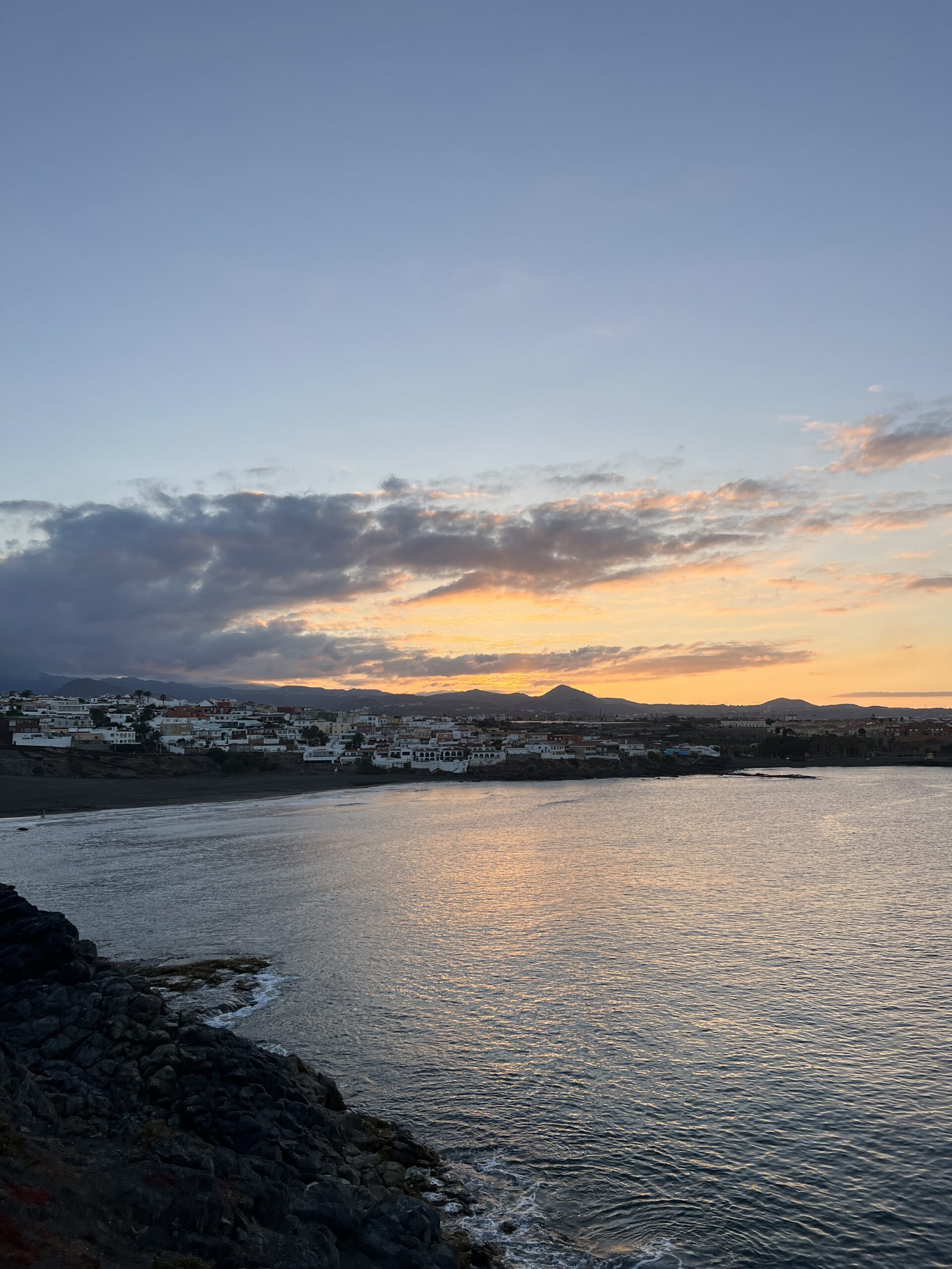 The Telde coastline glows at sunset, with whitewashed houses lining the shore and volcanic peaks rising beyond the bay.