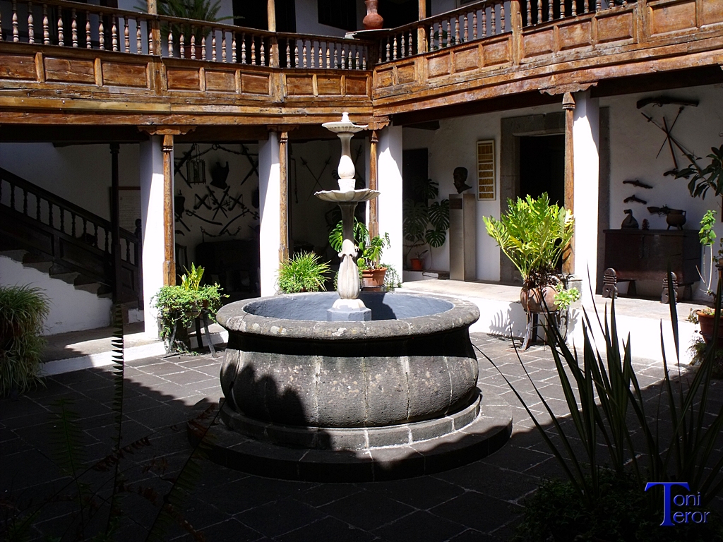 A traditional courtyard in Teror with a central stone well, lush potted plants and timber balconies typical of Canarian highland architecture.