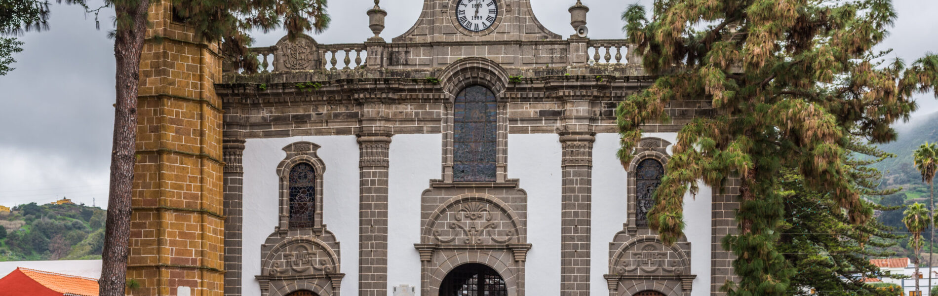 Stone church with white walls, basalt columns and bell gable framed by palm trees.