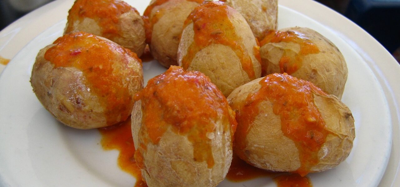 Eight golden-brown meatballs topped with red sauce on a white plate, with a blue-checked cloth and sauce bowl behind.