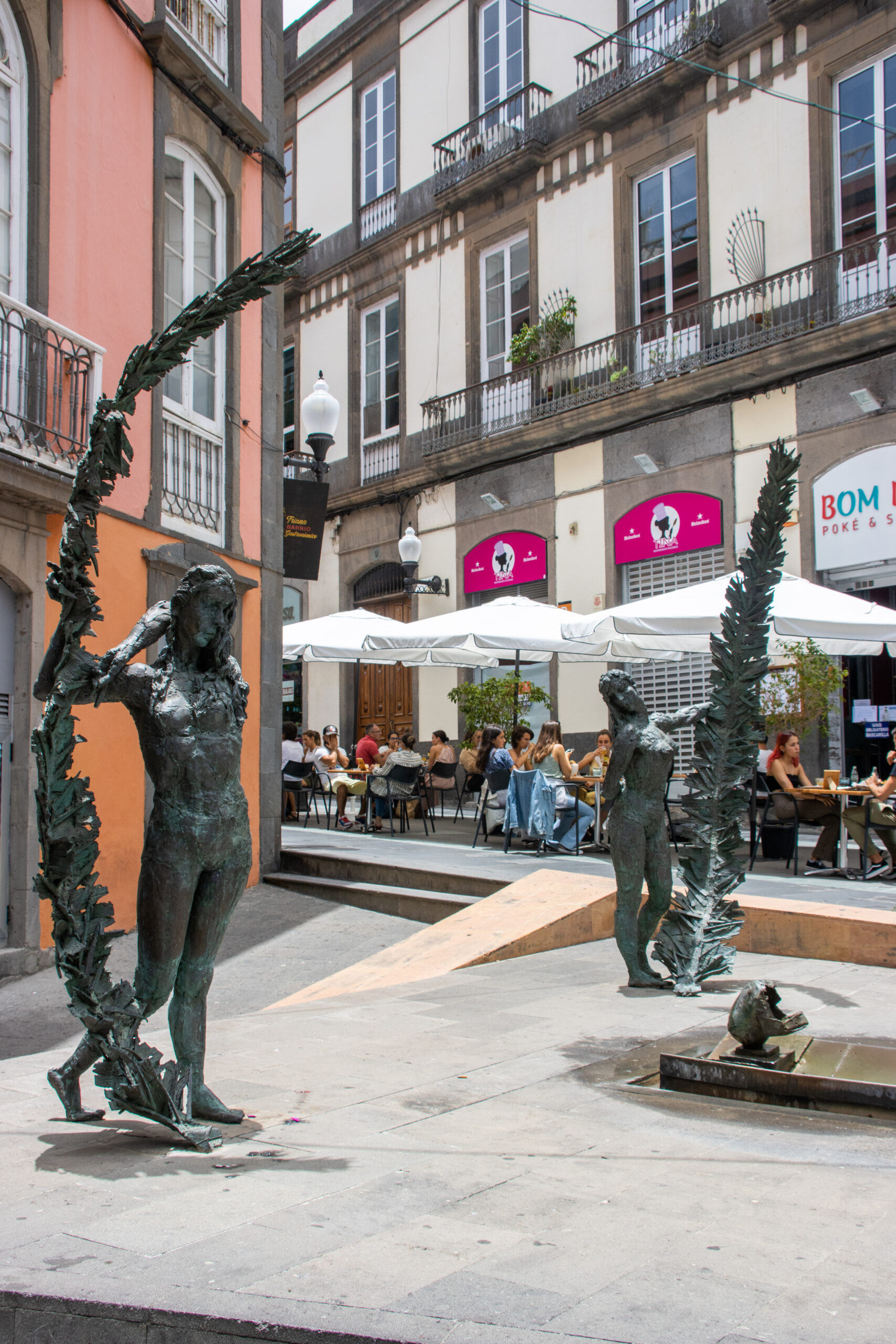 A public plaza in the Triana district features modern bronze sculptures of figures adorned with palm fronds, overlooked by historic colonial architecture and vibrant street-level cafés.