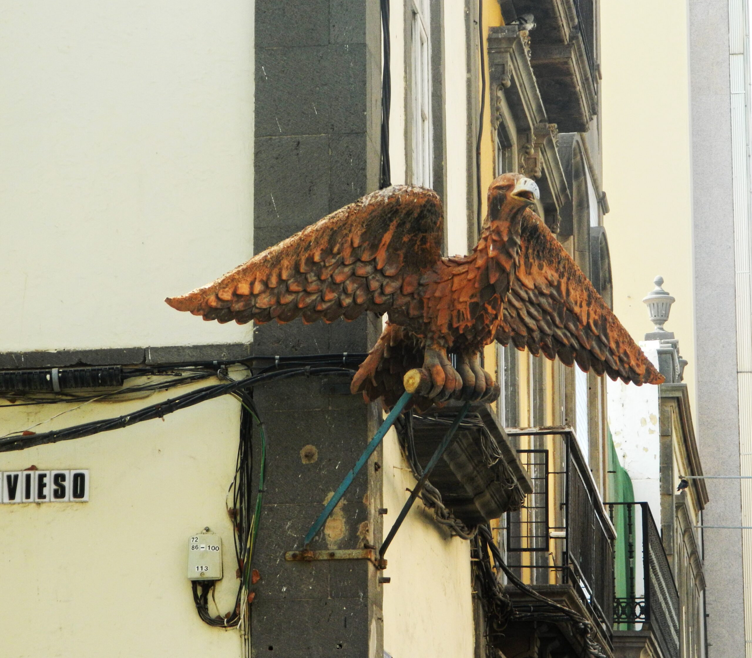 A weathered iron eagle perches on a building in the historic Triana district, its oxidized wings spread wide against the pale yellow facade.