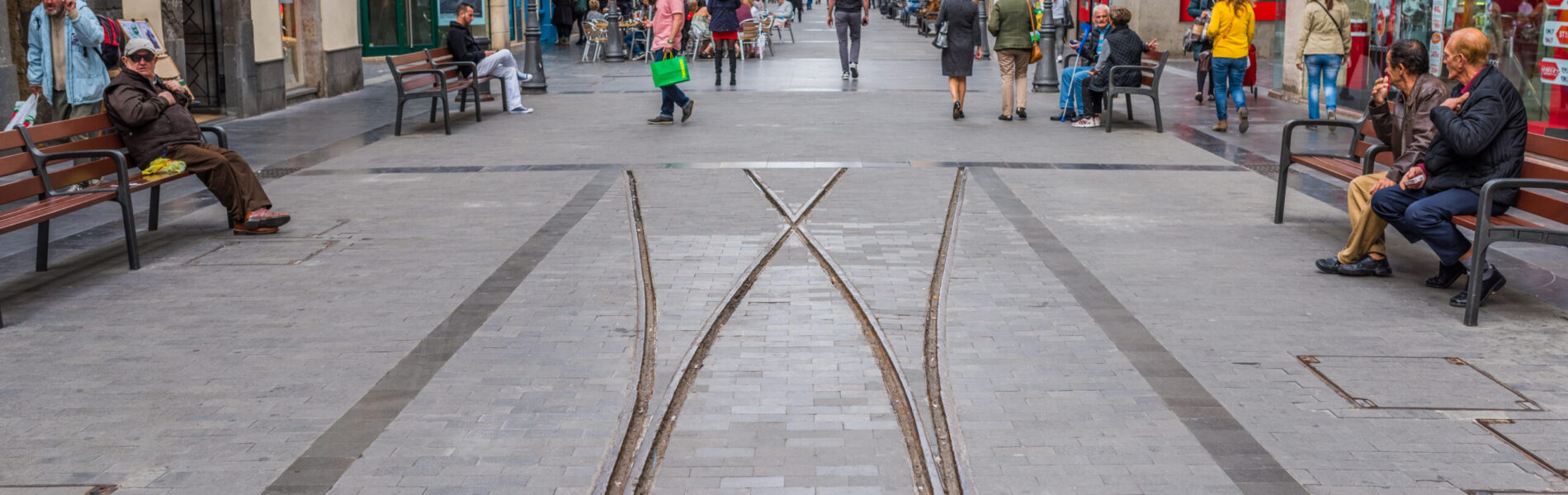 Pedestrian shopping street with embedded tram tracks, surrounded by historic buildings, shops, and people sitting on benches