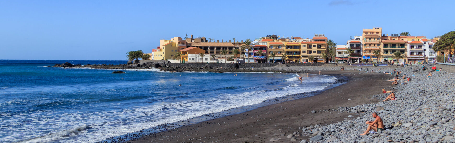 Dark sand beach with rocky foreground, blue Atlantic waves, and colorful apartment buildings lining the shore under clear sky