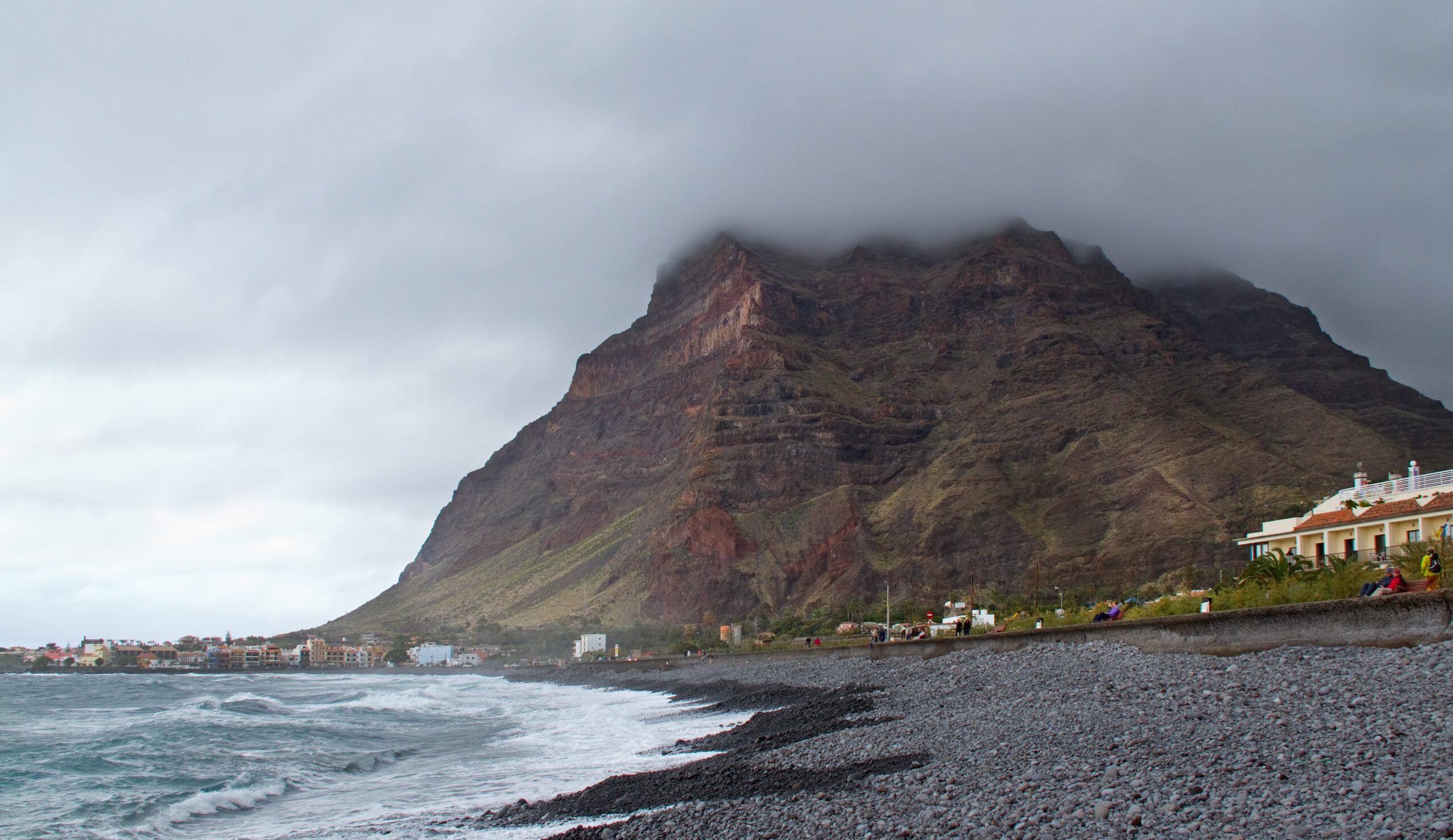 Valle Gran Rey's dramatic backdrop: a distinctive volcanic peak looms over the coastal settlement and pebble beach, with the Atlantic waves in the foreground.