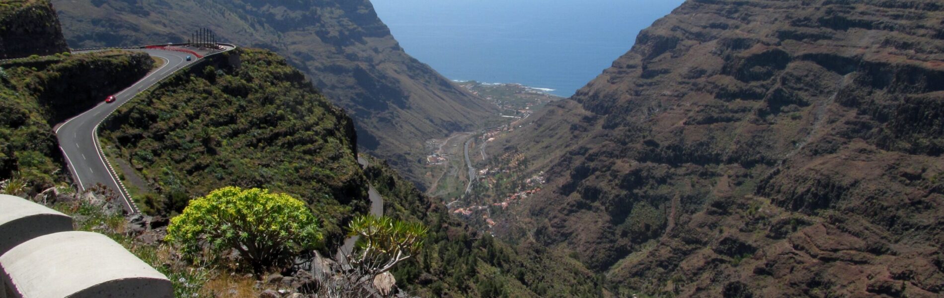 A winding road descends through a deep valley with steep volcanic slopes, ocean visible in the distance under blue sky.
