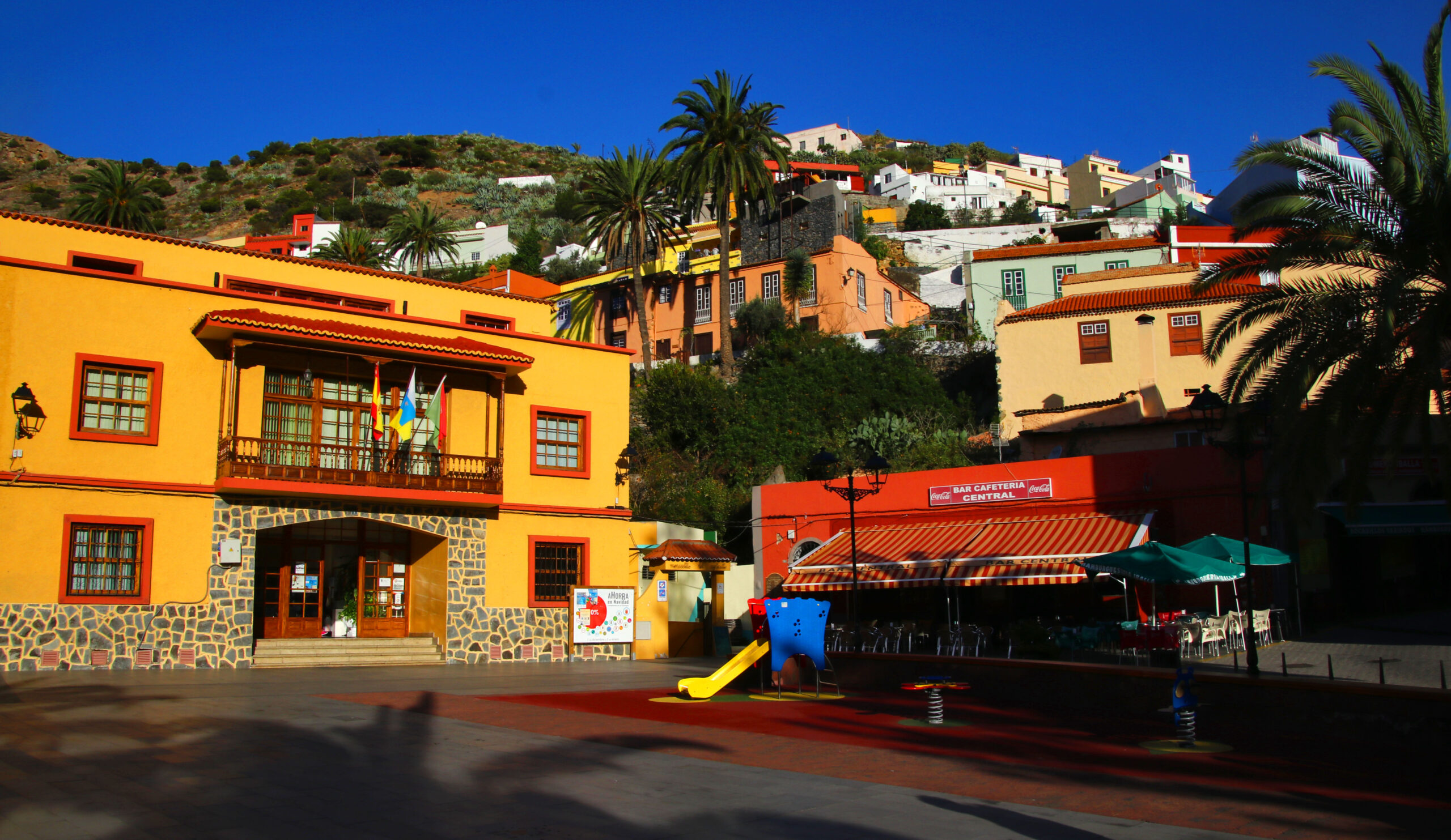 The charming main plaza of Vallehermoso features striking yellow and orange colonial-style buildings with local shops and cafés, set against the backdrop of whitewashed houses climbing the hillside.