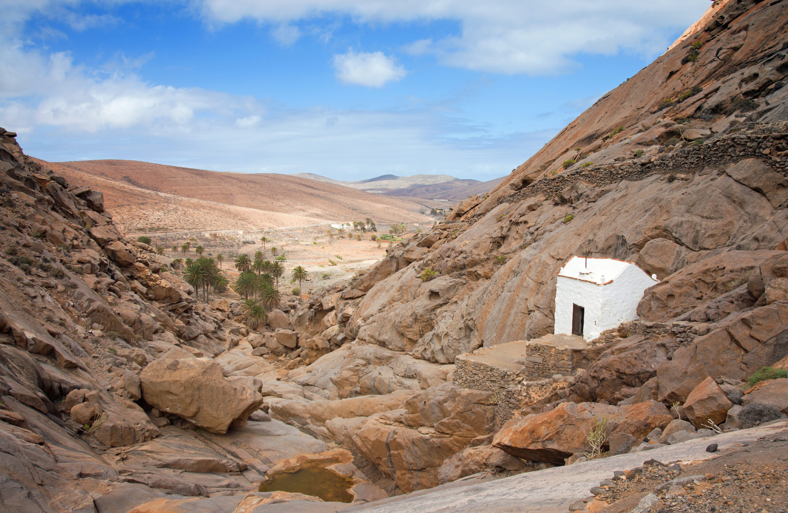 A whitewashed hermitage sits sheltered in the rocky ravine of Vega de Río Palmas, with palm trees dotting the arid valley floor beyond.