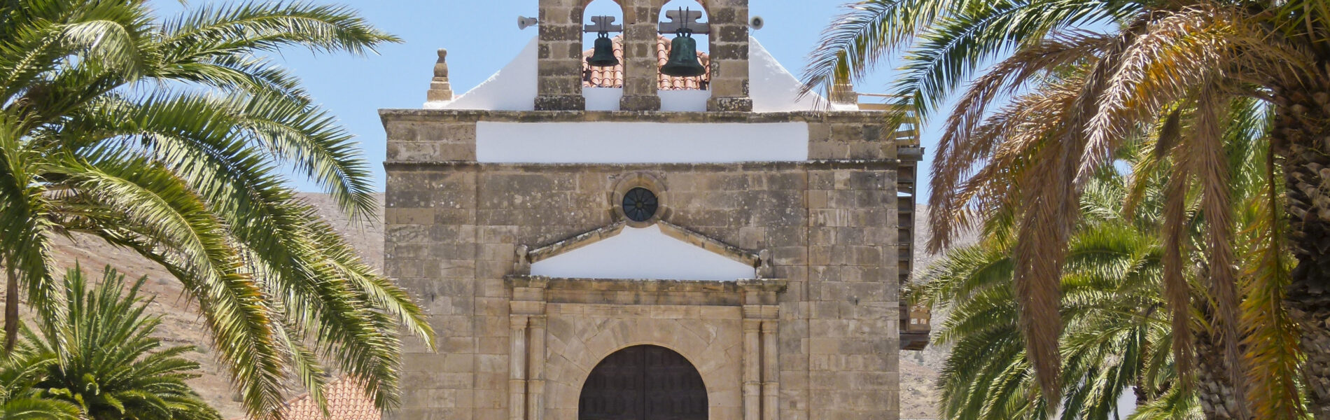 Stone chapel with bell tower framed by palm trees on a white paved plaza under clear sky
