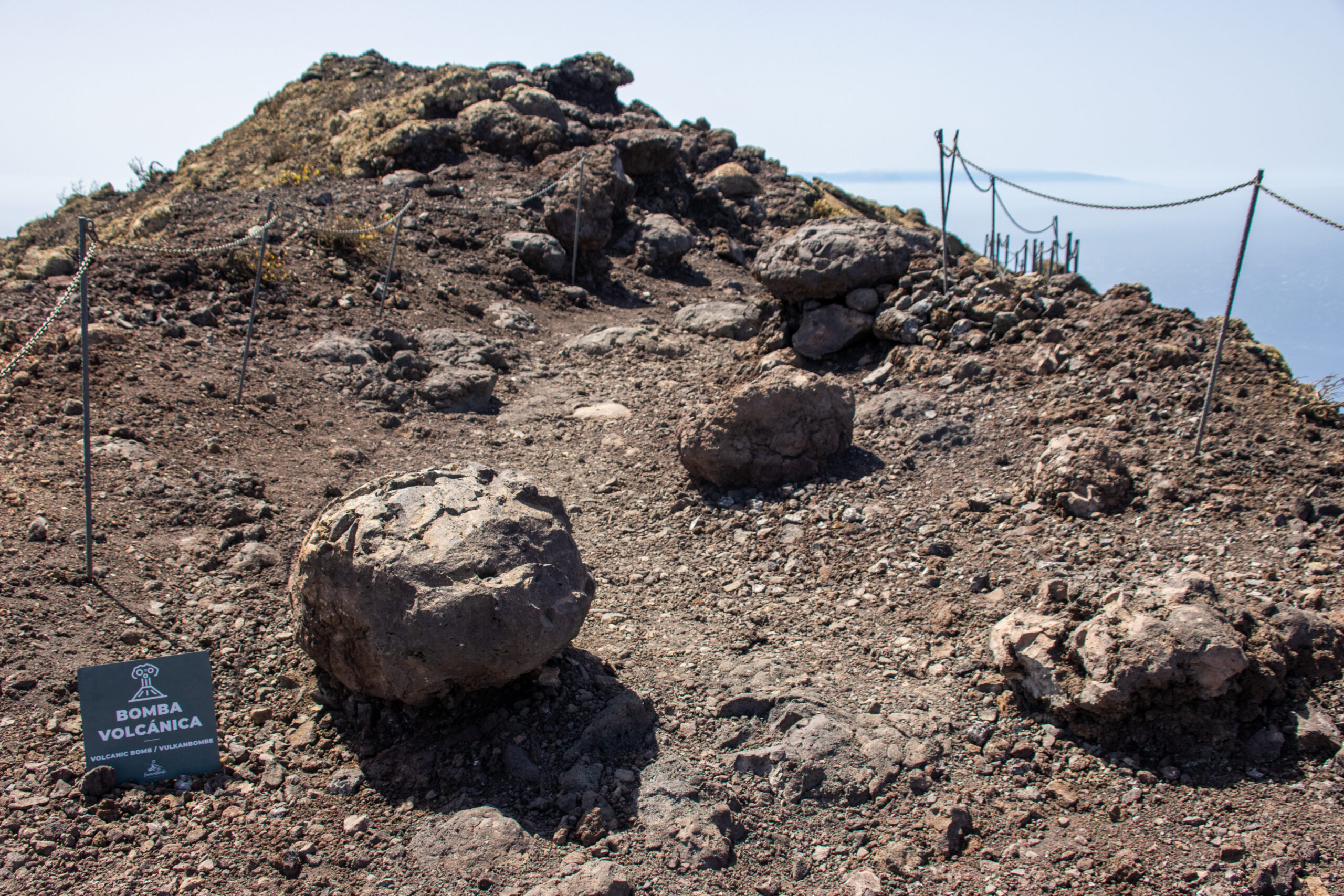 The volcanic landscape of San Antonio features characteristic lava bombs and loose volcanic material, with safety barriers and interpretive signage marking the crater slope.