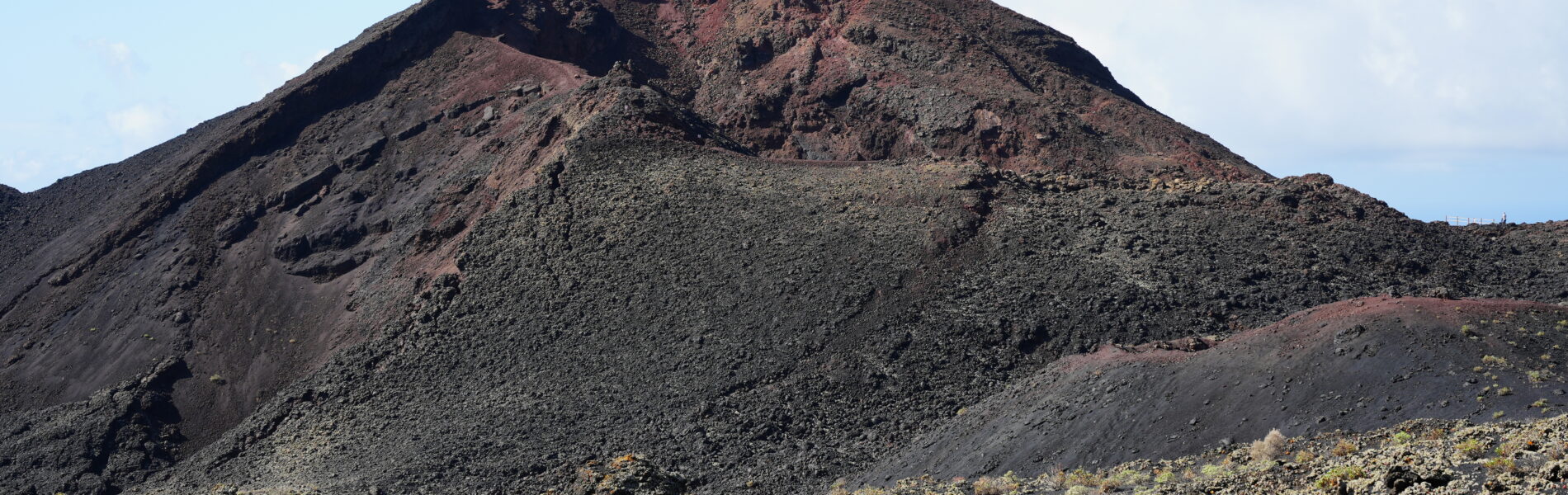 Dark volcanic cone with red-brown summit crater, surrounded by black and gray lava fields dotted with sparse yellow-green vegetation under blue sky