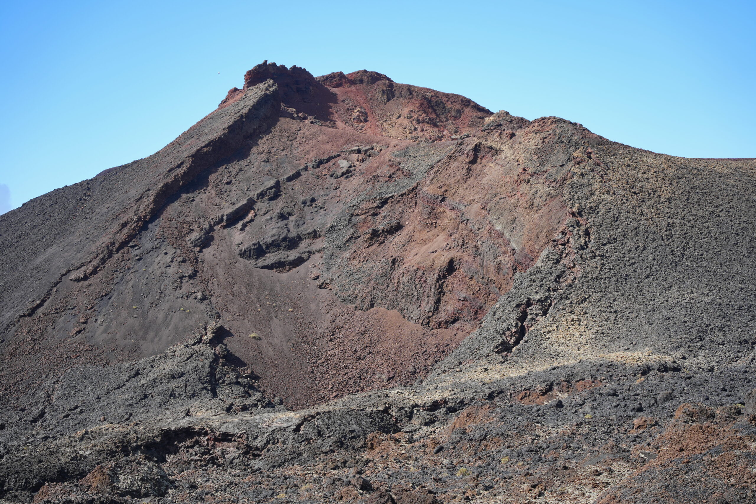 The stark volcanic cone of Teneguía displays layered deposits of dark lava and reddish oxidized rock, with loose scree covering its steep flanks.